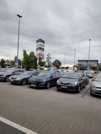 A medium-shot, eye-level, outdoor photograph captures a parking lot in front of the Galeria Wołomin shopping center, located in Wołomin, Poland, under an overcast sky. The parking lot is paved with interlocking gray concrete pavers, with white lines delineating parking spaces. In the foreground, several cars are parked, including a dark gray Toyota Avensis wagon with the license plate "WWY 72404" and a blue Renault Megane with the license plate "WY 8633J." To the left, a silver Mercedes-Benz CLA is partially visible.

In the mid-ground, a tall, multi-sided sign tower displays various store logos and names, including "GALERIA WOŁOMIN," "Carrefour," "LEROY MERLIN," "CCC," and "HELIOS." Several trees with green foliage are situated behind the sign tower and to the left, with residential buildings visible in the distance. Two blue parking signs are also visible.

The background features the modern facade of the Galeria Wołomin, with large windows and an entrance. A tall lamppost stands to the right of the shopping center. The sky is a uniform gray, suggesting cloudy weather. Several dark birds are visible flying in the sky. The overall scene depicts a typical day at a suburban shopping center.