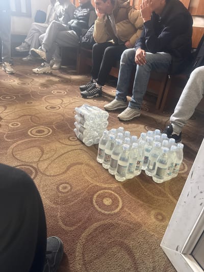The scene depicts a group of people sitting on chairs, seemingly indoors, with several stacks of bottled water on the floor between them. The floor is carpeted with a circular pattern design. The photo appears to be taken from a high-angle perspective, possibly by someone standing or sitting above the subjects. A small portion of a white object or surface is visible in the upper right corner. Without additional information, it's difficult to confirm that the location is in Charentsavan, Armenia.