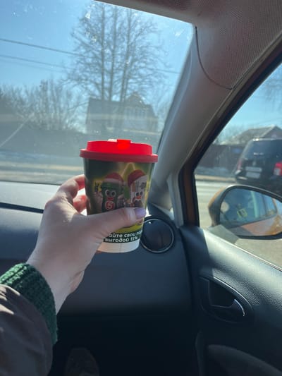 An image captured from the interior of a vehicle, in Yasenevo, Russia, displays a sunlit street scene. A person's right hand, with light skin tone and a light purple fingernail, holds a dark green disposable cup with a red lid in the foreground. The cup is adorned with cartoon figures, including a green character wearing a Santa hat and displaying a peace sign, marked with a white '5', and partially visible Cyrillic text on a white band: "айте свои пар выгодoй 15%". The individual wears a dark brown jacket with a green knitted cuff. The vehicle's dark grey dashboard and door panel are visible, along with an orange/brown side mirror reflecting a yellow object or sign marked '13'. Through the windshield, a clear blue sky with intense sunlight creates glare. Bare deciduous trees, overhead power lines, and a residential building with a pitched roof are visible in the background. A dark-colored SUV is parked or moving behind and to the right of the observer's vehicle.