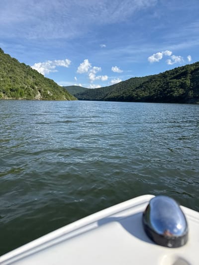 The image presents a view from a boat on a lake, bordered by a forested hillside under a sky with scattered clouds. The location is Dabovets, Bulgaria. The right side of the frame shows a portion of the boat, including a white railing and a small navigation light. The lake occupies the central part of the image, with the water surface showing slight ripples. On the left, a steep hill is covered in dense green vegetation. The sky above features patches of blue interspersed with white clouds. There are no people visible in the image.