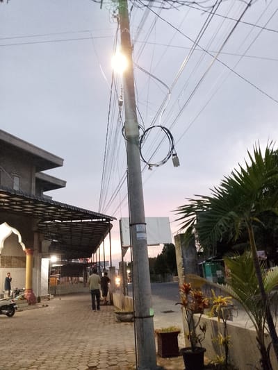 The image depicts a street scene in Geulanggang Teungoh, Indonesia. The time appears to be dusk, with the sky exhibiting a soft gradient of light blue and pink hues. A tall, concrete utility pole dominates the foreground, heavily laden with a complex network of electrical wires. One of these wires is coiled around the pole. A bright light fixture is attached to the pole, emitting a strong glare.

To the left of the pole, a modern building with a flat roof and angular architecture is visible. A covered walkway with a checkered roof extends from this building, creating a shaded area. Several people are present. One individual, wearing a dark jacket and light-colored pants, stands near a motorcycle parked under the covered walkway. Further down the walkway, two men are walking away from the camera. One is wearing a light-colored shirt and dark pants, while the other is in a dark shirt and shorts.

On the right side of the image, a palm tree with distinct fronds stands tall. In front of it, two potted plants with red and yellow foliage are visible. The ground is paved with interlocking brick pavers. In the background, a street stretches away, with buildings and trees lining its sides. A small, white sign is affixed to the utility pole. The overall impression is a quiet, urban street scene as evening approaches.