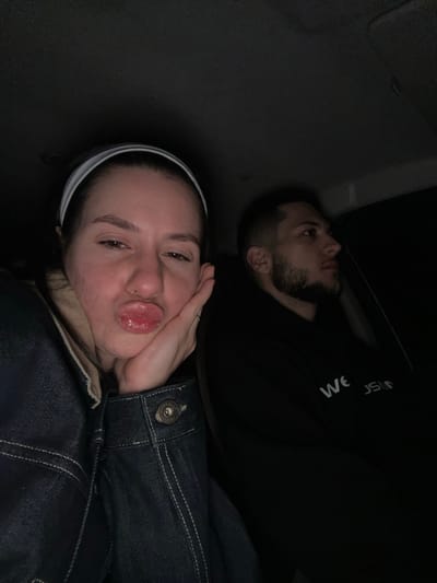 The image shows two people lying down. A woman is in the foreground, wearing a denim jacket and a white headband, making a pouting face. A man with a beard is lying behind her, wearing a black top with white lettering. The setting appears to be inside a vehicle, possibly a car. The background is dark and indistinct. The location in the city of Yastrebovo, Bulgaria is not discernable.