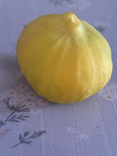 The image shows a single, ripe, yellow fig placed on a light-colored patterned surface. The surface appears to be a tablecloth or placemat with a subtle floral design and scattered small yellow dots. The fig is oval-shaped with a slightly irregular top, exhibiting subtle ridges and color variations, with some areas appearing lighter yellow or greenish. The background is out of focus and appears to be a solid, muted color. There is no indication of a city or specific location within Tasbuget, Kazakhstan, in this image.