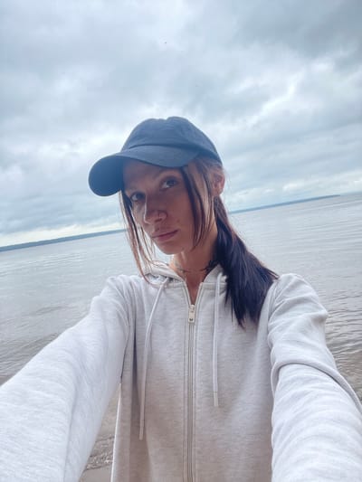A young woman wearing a black baseball cap and a light gray zip-up hoodie is taking a selfie outdoors. She has long dark hair pulled back into a ponytail, and a nose ring and lip ring. She is looking directly at the camera with a neutral expression. The background shows a body of water under a cloudy sky, suggesting a cool or overcast day. The location is identified as Ikskoe Ust'e, Russia, which is a coastal area or situated near a large body of water.