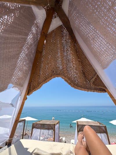 The image captures a first-person perspective from within a rustic beach cabana, providing a view of a serene seaside scene in Vrba, Montenegro. In the immediate foreground, the viewer's tanned bare legs and feet rest on a light-colored cushioned lounger, extending towards the lower right. Overhead, the cabana's roof is constructed from a dark brown, loosely woven natural fiber material, with sections of translucent white mesh fabric interwoven, allowing sunlight to filter through and create dappled light patterns.
Beyond the cabana, the midground reveals a pebbly beach populated with other wooden cabanas featuring white sheer curtains and white beach umbrellas. Several individuals are visible on the beach and wading in the clear, blue-green water near the shore. A distinct line of white buoys or markers stretches horizontally across the ocean, delineating a swimming area. In the background, the calm, expansive sea extends to meet a bright, cloudless blue sky at the horizon. The scene depicts a sunny and tranquil day at the beach.