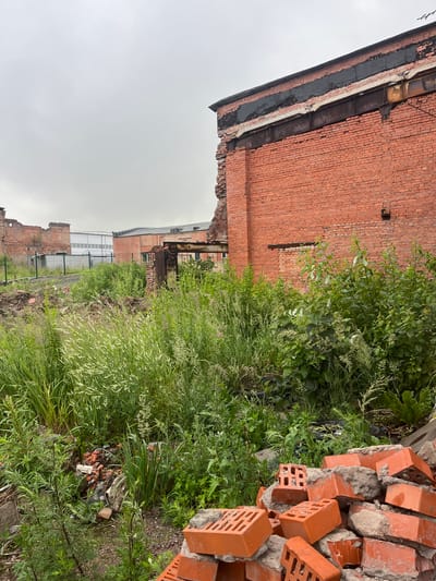 This media file depicts an outdoor scene in what appears to be an industrial or abandoned area, likely in Imeni Sverdlova, Russia. The dominant subjects are a dilapidated brick building and a pile of construction debris, primarily consisting of broken red bricks and mortar, along with some scattered vegetation.
The brick building shows signs of damage and disrepair, with sections of the wall missing and the roofline appearing weathered. Adjacent to the building, a dense growth of green vegetation, including tall grasses and leafy plants, encroaches on the scene. In the foreground, a large pile of orange-red bricks, some intact and others broken, is piled up haphazardly. Mixed within the vegetation and debris are also some pieces of what look like scrap metal and other discarded materials.
The lighting suggests it is daytime, with an overcast sky that casts a muted, diffused light over the scene, hinting at cloudy weather. There are no people visible in the media. The overall impression is one of decay and abandonment, with nature reclaiming the man-made structures. The location cues of brick construction and industrial elements are consistent with the provided location context. No visible text is present in the image.
