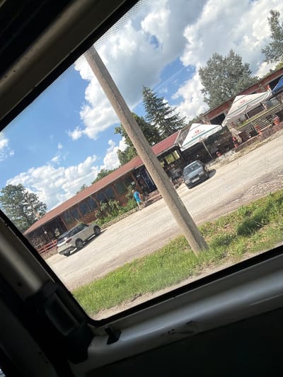 A low-angle shot taken from inside a vehicle shows a scene outside a roadside establishment in Mikre, Bulgaria. A long, rustic wooden building with a reddish-brown roof and numerous windows runs parallel to a paved road. In front of the building, there are several white umbrellas with the text "Kamenitza" visible, suggesting it is a restaurant or beer garden. A beige SUV is parked on the asphalt in front of the building, and a dark gray sedan is further down the road. A single person wearing a blue shirt and shorts is standing near the entrance of the building. A thick, gray concrete pole stands prominently in the foreground, obscuring part of the view. The sky above is blue with scattered white clouds, and trees are visible behind the building and along the roadside, indicating a rural or semi-rural setting. The road itself appears to be in moderate condition with patches of grass and dirt along its edges.