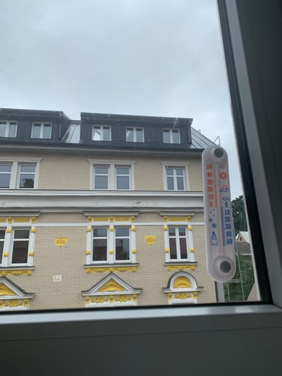 This image shows a thermometer outside a building, viewed through a window. The thermometer is white with red and blue markings. The blue markings indicate temperatures from 0 to 50 degrees Celsius, with icons representing snow, a snowflake, and a snowman at the lower end, and clouds and rain at the higher end. The red markings indicate temperatures from 0 to 50 degrees Celsius, with icons representing a sun and a sun with clouds at the higher end, and a thermometer with a downward pointing red line at the lower end. The red liquid in the thermometer appears to be at approximately 10 degrees Celsius.
The building behind the thermometer is multi-story with light beige brickwork and white-framed windows. Some windows have decorative gold elements above them, resembling stylized sunbursts or crests. The building also has a darker facade on the left side, with windows and what appear to be gutters.
The weather appears overcast and possibly damp, as indicated by the thermometer's reading and the general brightness of the sky. The time of day is difficult to ascertain definitively, but the overcast conditions suggest it is likely daytime. The image is taken from inside a room, looking out through a window frame, which is visible in the foreground and on the right side of the image. The specific location of Vejprty, Czechia, is provided as context. No people are visible in the image.