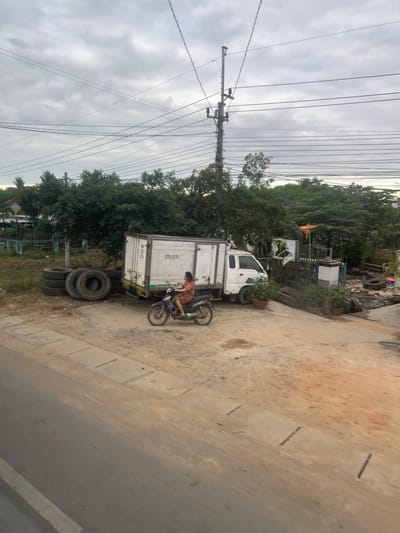 An outdoor, daytime scene in Ninh Tây, Vietnam, captured under an overcast sky. The foreground features a paved road with a concrete curb separating it from an expansive, uneven dirt and gravel area. In the midground, a woman in an orange patterned dress is seated on a dark gray scooter, oriented right, with her right foot resting on the ground, indicating a stationary or slow-moving posture. To the left of the scooter, a stack of five large, dark vehicle tires is present. Behind the woman and the tire stack, a white box truck, marked with "57S-3179" on its side, is parked. The truck's front cab is white. To the right of the truck, an unpaved incline leads upward past a low dark fence and a decorative pillar, revealing additional tires, discarded materials, and potted plants. The background is dominated by dense, lush green foliage and trees, partially obscuring what appear to be residential structures on the far left. A tall utility pole with multiple intersecting overhead power lines extends vertically into the gray, cloud-covered sky.