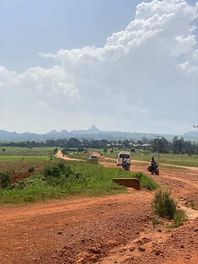 An eye-level, long shot captures a dusty, red dirt road winding through a rural landscape under a bright, cloudy sky. The road is the central focus, with its reddish-brown surface marked by tire tracks and scattered pebbles. On the right side of the road, a small, green bush grows from the embankment.

Several vehicles are present on the road. In the foreground, on the right, a motorcycle carries a rider and a substantial amount of luggage. Further down the road, another motorcycle with two riders is approaching. A white van is also visible on the road, followed by a dark-colored car. In the distance, a few more vehicles are scattered along the winding path.

The road is flanked by fields of lush green grass and some patches of cultivated land. In the mid-ground, a line of trees separates the fields, and beyond them, a scattered collection of buildings suggests a small village or settlement. The background is dominated by a hazy mountain range with distinctive rocky formations, silhouetted against the sky. The sky itself is a pale blue, with large, white cumulus clouds dominating the upper portion of the image. The overall impression is one of a remote, possibly developing, rural area. The image does not depict the city of Rim, Nigeria, as there is no indication of its location.