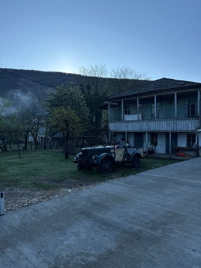 The image depicts a two-story wooden building in Gachedili, Georgia, situated next to a concrete road. A vintage tractor is parked on the grassy area in front of the building. Trees and a mountain are visible in the background, and there is smoke or mist rising in the lower left. The sky is clear and blue.
