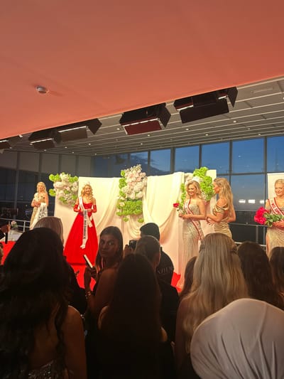 The image depicts a Miss Denmark beauty pageant event taking place in Skovshoved, Denmark. Several contestants wearing sashes with titles such as "FINALIST - MISS DANMARK 26" and "MISS SUPR" are visible on a stage decorated with white fabric and floral arrangements. They are holding bouquets of flowers. A crowd of people, primarily women, is observing the event and taking pictures with their phones and cameras. The background shows a multi-story building with large windows, suggesting an indoor venue with outside views. The wall is light pink.