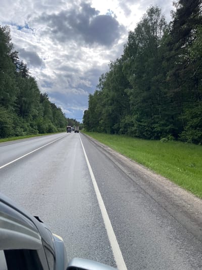 The image shows a straight asphalt road with white lines marking the lanes. Dense green trees line both sides of the road. In the distance, there are two trucks driving away. The sky above is filled with clouds. The picture appears to be taken from inside a moving car, as part of the car's roof and side mirror are visible in the lower right corner. The location is likely the road leading to or passing through Semyonovskoye, Russia given that the image was specified to be in that city.