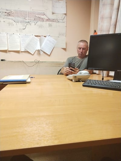 A middle-aged man, with short grey hair and a gray t-shirt, is seated at a wooden desk, looking down at his smartphone. He is holding the phone in his right hand and appears to be engaged with it. In front of him is a white landline telephone and a black computer keyboard. To his left, on the desk, are two stacked folders, one blue and one white, with a blue pen on top. Behind him, a large schematic diagram of railway tracks is pinned to the wall, along with several loose documents. To the right of the man, a computer monitor displays a black screen. The room has pale peach walls and checkered curtains are visible on the right side of the frame. The image appears to be taken in an office environment.