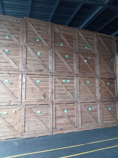 A high-angle, eye-level shot captures a vast wall of stacked wooden crates within a covered outdoor area. The crates are uniformly constructed with horizontal planking and diagonal bracing, creating a textured, geometric pattern. Each crate appears to be roughly square and is stacked several levels high. Small, square, green and white stickers are affixed to many of the crates, some of which have legible text or barcodes. The background reveals the underside of a corrugated metal roof supported by dark metal beams, suggesting a warehouse or covered storage facility. The foreground shows a dark asphalt surface with two parallel yellow lines, indicating a road or walkway. There are no people or animals visible in the scene, and the environment is clean and orderly. The image appears to be taken in Biloziria, Ukraine, in a storage facility or logistics hub.