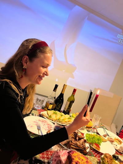 A woman with a maroon headband smiles, holding a phone to capture an image of a lit candle. A table laden with food and beverages is in view. Wine bottles stand near plates, and a projection screen behind suggests an event or gathering. The setting indicates a festive occasion with a celebratory atmosphere.
