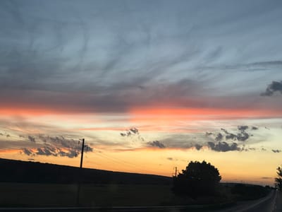 This is a photograph taken from a moving vehicle, likely on a road, looking towards a sunset. The sky is filled with clouds displaying a gradient of colors from a pale blue at the top to vibrant orange and pink hues near the horizon. In the foreground, a dark silhouette of a hilly landscape with trees and vegetation is visible. A utility pole with power lines stretches across the scene from left to right, a prominent feature against the colorful sky. The road itself is also silhouetted, suggesting it is a dark asphalt surface. The image captures the natural beauty of a sunset over a rural or semi-rural area, potentially in or near Veselina, Bulgaria.