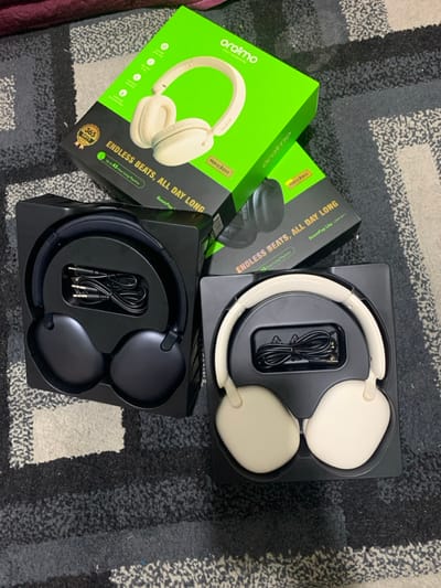 The image showcases two sets of over-ear headphones. One set is black and the other is cream-colored. Both sets are presented in their original black packaging, with their respective cables neatly stored within. Beside the cream headphones is their box, a bright green, angled to show the brand name "oraimo" and the phrase "ENDLESS BEATS, ALL DAY LONG." A second box is also partially visible, featuring the black headphones. The scene is set against a gray and black patterned carpet.