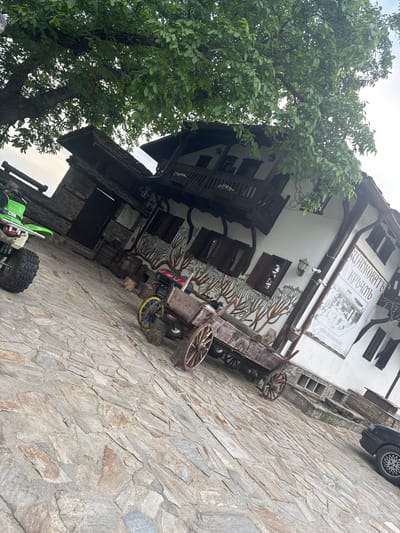 This is a photograph of a building with an old-fashioned, rustic architectural style, likely a restaurant or guesthouse, located in Delchevo, Bulgaria. The building has multiple stories with wooden balconies and windows. The facade is adorned with painted tree motifs and a large sign with text that appears to be in Cyrillic.
In front of the building, on a cobblestone pathway, is a decorative, wooden cart with large, spoked wheels. A bright green, child-sized ATV or go-kart is parked on the pathway in the lower left portion of the image. A large tree with dense green foliage is prominently featured to the left of the building, partially obscuring the upper floors.
The scene appears to be outdoors, with an overcast sky suggesting it might be a cloudy day, possibly in the late morning or afternoon. There are no people visible in the image, and the overall atmosphere is still and unpopulated. The combination of the building's design, the wooden cart, and the natural elements like the tree suggests a setting that emphasizes traditional or rural aesthetics.
Visible text includes "DELCHEVSKO.EU" and a phone number "0893 212 558" on the large sign. There is also some Cyrillic text on the sign, which likely identifies the establishment. The presence of a modern vehicle (the ATV) and a car partially visible at the top right contrasts with the older elements of the scene.