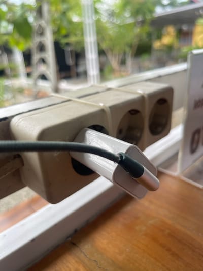 In Pulokiton, Indonesia, a white electrical extension cord with two outlets is secured with two white zip ties to a white horizontal bar. A white phone charger is plugged into the leftmost outlet, with a dark green cable extending from it. The background is blurred, showing greenery and a building, suggesting an outdoor or semi-outdoor setting. The image is taken from a slightly elevated angle, focusing on the electrical outlet and charger.