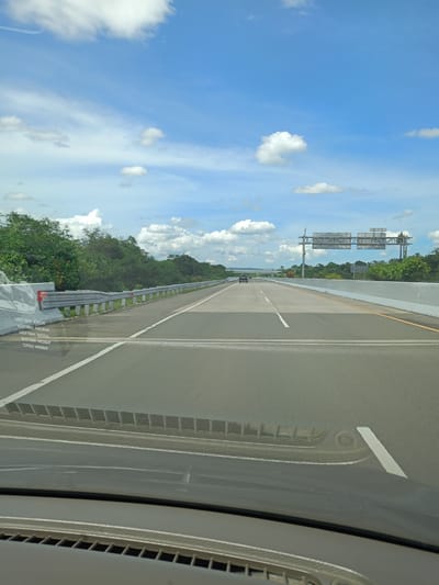 The image depicts a view from inside a vehicle traveling on a highway in what appears to be the Seulimeum area of Indonesia. The highway is multi-lane, with concrete barriers on the right and guardrails on the left. Vegetation lines both sides of the highway. A sign structure spans the highway further ahead. A single dark-colored vehicle is visible in the distance on the same side. The sky is blue with scattered white clouds. The car's dashboard is visible in the foreground.
