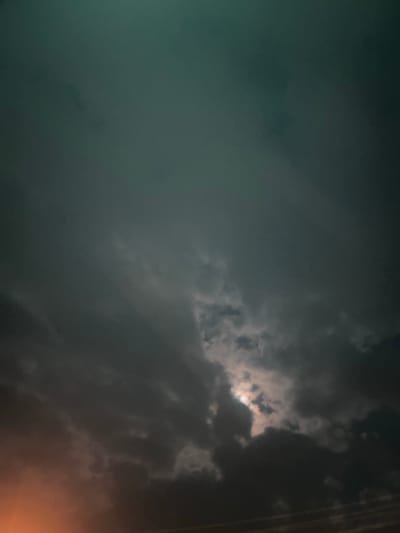 The image shows a cloudy sky with the moon partially visible through a break in the clouds. The sky is dark and appears to be experiencing stormy weather, with dark, heavy clouds dominating the scene. There is a bright orange glow emanating from the lower left corner of the image, likely from a light source on the horizon. The overall atmosphere is dramatic and moody. The image does not provide any specific details or context related to the city of Белозерки, Russia.