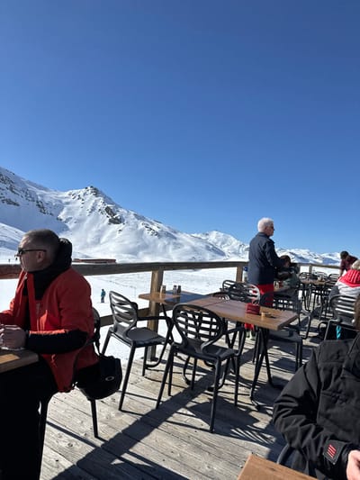 The image shows a mountainside view from a wooden deck in the French Alps. The location is Les Belleville, France, at coordinates 45.281, 6.573. Several people are seated at a table, some are facing a snow-covered mountain range. The table is made of wood and has various objects on it, including drinks and snacks. The deck is made of wood planks and has black metal chairs with a unique open design. The deck is partially shaded, casting shadows onto the wooden surface. A person in red is sitting at a table near the bottom of the image and is looking at their hands while the person at the top of the image is looking directly at the snow covered mountains. The sky is a clear, bright blue, and the snow is white. The scene is calm and peaceful, with a relaxed atmosphere. The people appear to be enjoying the beautiful views and the good weather. The scene also shows a ski lift to the left of the frame.