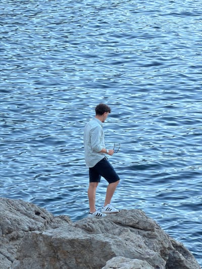 This is a vertically oriented photograph of a person on a rocky cliff overlooking a body of water. The person is a young man with curly brown hair. He is wearing a light-colored long-sleeved shirt, dark shorts, and white sneakers with black stripes. He is holding a pair of glasses in his right hand.

The man is positioned on the edge of a textured, rocky cliff face. His body is mostly horizontal in the frame, with his legs extending towards the bottom left and his torso angled towards the top left. He is looking to his left, away from the viewer. The cliff itself is a prominent feature, with rough surfaces and visible cracks and crevices.

To the left of the cliff, a vast expanse of rippling blue water fills the majority of the frame. The water's surface is characterized by numerous highlights and shadows, creating a dynamic and textured appearance. The lighting suggests it is daytime, with bright illumination that casts sharp contrasts on the water and the rock. The scene evokes a sense of solitude and perhaps contemplation, as the man appears to be observing the water. There is no visible text in the image.