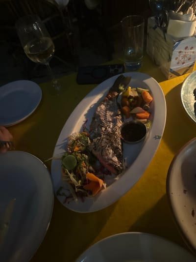 A white platter presents a cooked fish, accompanied by a small dish of dark sauce, roasted vegetables, and a salad with a lime wedge. Surrounding the platter on a yellow surface are multiple plates, a wine glass, a tumbler, a phone, napkins, and silverware. Part of a hand holding cutlery is visible at the bottom of the frame. The lighting suggests an evening setting.