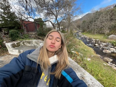 The image shows a young woman lying on a concrete surface with a stream and trees in the background. She is wearing a dark blue and black jacket with a grey hood. Her eyes are closed and her lips are slightly parted, suggesting a relaxed or peaceful state. Her blonde hair is spread out around her head.

The scene appears to be outdoors, possibly in a natural setting given the presence of the stream, rocks, and trees. The foliage on the trees suggests it might be late autumn or early winter, as many branches are bare. The sky is visible and appears to be a clear blue. There is a red-roofed building in the background, possibly a house or cabin, partially obscured by trees and greenery.

The perspective of the photo is unusual, as the camera seems to be positioned very close to the woman's face, and the entire image is rotated. It appears to be a self-portrait taken by the woman herself.

There is no visible text in the image. The overall mood of the image is serene and contemplative, with the natural environment providing a tranquil backdrop. The lighting suggests it is daytime, likely mid-morning or afternoon based on the brightness.