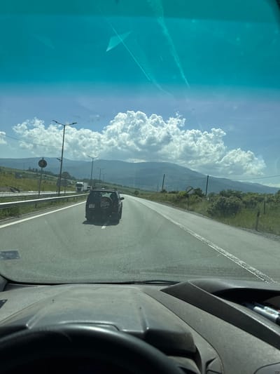 The image shows a view from the inside of a vehicle, looking forward through the windshield. The vehicle is on a paved road with two lanes in the visible direction of travel. A dark-colored SUV with a spare tire mounted on the back is in front of the vehicle, in the right lane. The license plate of the SUV is partially visible and appears to be black with white lettering. The road is bordered by a metal guardrail on the left and grassy slopes with some vegetation on the right.
In the distance, there are mountains under a bright blue sky with large, fluffy white clouds. Several lampposts are visible along the roadside. There are also some signs, including a circular sign with a diagonal red line on a white background, indicating a no-entry zone or similar restriction. In the far distance, a white truck is visible.
The foreground of the image shows the dashboard of the vehicle, including a portion of the steering wheel. The dashboard and windshield appear to have some dust or dirt on them, possibly from the environment. The overall scene suggests a daytime journey on a highway or main road, likely in a rural or mountainous area. The location is identified as the city of Golemo Buchino, Bulgaria.