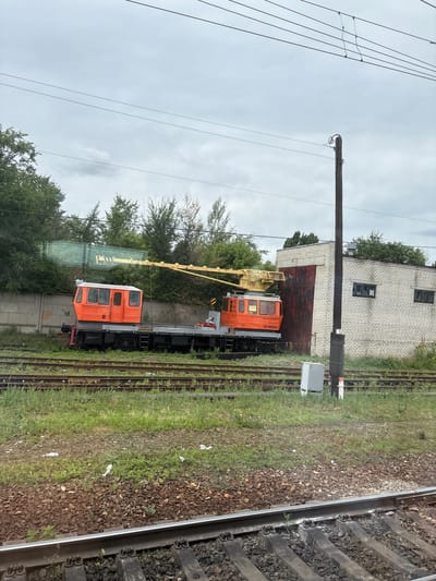 In Povorino, Russia, an outdoor scene depicts a train yard under an overcast sky. Two orange train cars, identified as cranes, are positioned on railway tracks. The crane on the left is closer to the foreground and is partially obscured by trees and a concrete wall. The crane on the right is more visible, featuring a yellow boom and the number "18100024" on its side. Adjacent to the tracks is a white brick building with a large, rusted metal door and two dark-paned windows. A tall, dark utility pole stands near the building. The foreground shows multiple sets of railroad tracks running parallel, with ballast and grass between them. Scattered litter is visible on the ground. Trees and foliage line the background. The overall atmosphere suggests an industrial or maintenance area within the city.