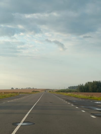 A long, straight asphalt road stretches into the distance under a cloudy sky. The road has a white solid line on the left and dashed white lines on the right, indicating multiple lanes. There are some small puddles of water on the right side of the road. Fields of dry grass and some trees line the sides of the road. In the distance, a single dark car is visible on the road. The scene appears to be in a rural area in the city of Chervonnoslobodsky Selsoviet, Belarus.