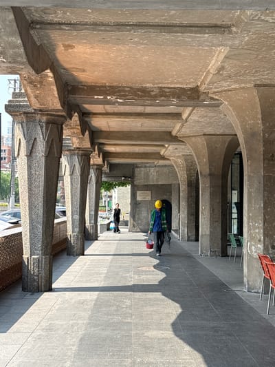The image captures a long, covered walkway supported by a series of stone columns and arches, giving it a tunnel-like appearance. The location is the Hongkou District in China. The columns are textured and feature decorative stepped detailing near the top. The arches above are made of a smoother, weathered stone. The walkway is paved with gray tiles, and shadows cast by the arches create a pattern along the ground and wall.
Two people are visible in the walkway. One is wearing a yellow hardhat, blue jacket, and carrying what appears to be equipment or supplies, including a red bucket. The second person is in the distance wearing darker clothing. They are walking away from the foreground.
In the background, there is an opening leading to an outdoor scene with greenery and buildings. Some modern amenities like a parked car and residential structures are visible in the distance. The scene suggests a juxtaposition of historical architecture with contemporary urban elements.