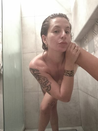 A wet, fair-skinned woman with wet hair and tattoos on her left arm and leg stands in a shower. She wears earrings and a ring on her left ring finger. The shower has light gray tiled walls and a partial glass door on the left. The woman is looking towards the camera and resting her right hand on her face. The image does not contain any information about the location of Inriville, Argentina.