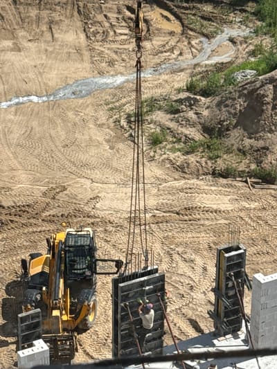 This aerial, top-down view captures a construction site in Mikołajki, Poland. The scene is dominated by a sandy, uneven terrain with numerous tire tracks indicating heavy machinery activity. A bright yellow excavator is parked on the sand, its boom arm extended. In the background, several large concrete formworks, reinforced with rebar, are visible. One worker, wearing a light-colored shirt and a cap, is actively engaged with the formwork, possibly preparing it for concrete pouring. The lighting suggests it's daytime, with distinct shadows cast by the machinery and structures. A thin, white stream, possibly water or a cement mixture, flows down a slight incline on the left side of the frame, cutting through the sandy ground. The overall impression is one of ongoing construction work under clear daylight conditions. No visible text is present in the image.