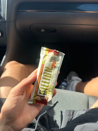 A person's hand holds a protein bar with "ONE IN A BILLION" printed on it, in front of a car's interior. The background includes car seats, a person's legs wearing athletic shorts and shoes, and a water bottle. The scene appears to be inside a car, possibly in Alès, France, given the text on the bar.