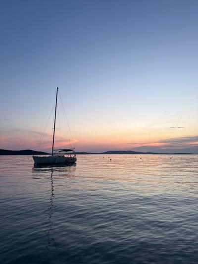 A sailboat named "DAZZLE DAZZLE" is anchored on calm water under a twilight sky. The sky transitions from a pale blue at the top to soft pinks and oranges near the horizon, where a silhouetted coastline with hills is visible. The water reflects the colors of the sky and the sailboat's mast. The scene appears to be in the city of Šibenik, Croatia, likely in a bay or harbor area. There are no people visible in the image.