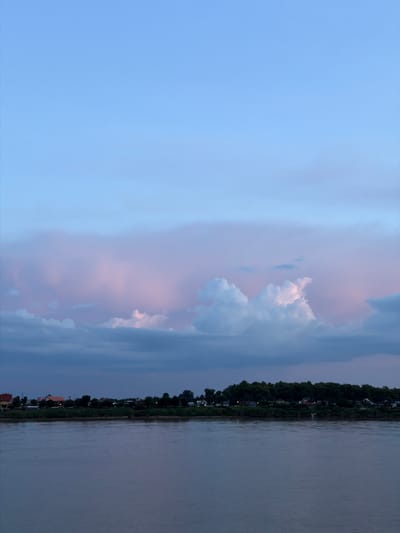 This image captures a serene twilight scene over the Mekong River, likely near Vientiane Capital, Laos. The upper portion of the frame is dominated by a vast, pale blue sky transitioning into softer hues of pink and lavender, indicative of sunset or sunrise. Wispy clouds are scattered across the sky, some tinged with the warm colors of the fading light, while others are a deeper, shadowed blue.

Below the sky, a line of lush green trees forms a dense border along the distant riverbank. Peeking through the foliage are several structures, including buildings with orange and white roofs, suggesting a small settlement or village nestled along the water. The lights from some of these buildings are faintly visible, twinkling against the darkening landscape.

The lower half of the image is occupied by the Mekong River itself. The water is calm and reflects the muted colors of the sky, appearing as a broad expanse of grayish-blue. The surface of the river shows subtle ripples, suggesting a gentle flow. There are no boats or people visible in the scene, contributing to the overall impression of tranquility and stillness. The image is framed vertically, emphasizing the expansiveness of the sky and river.