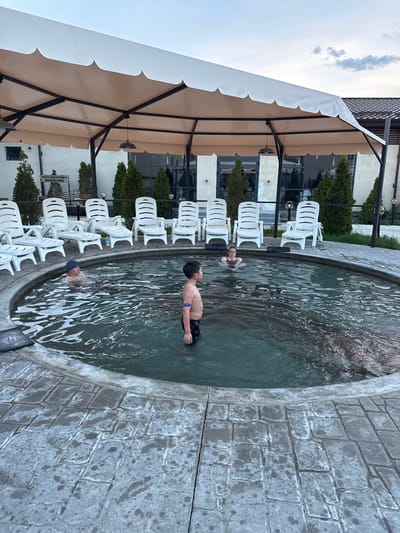 Here's a description of the image:

The photograph appears to be taken outdoors at a resort or recreational facility, potentially in Kara-Oy, Kyrgyzstan, given the location mentioned. A circular pool dominates the right side of the frame, containing three people: a boy standing up, and two others in the water. The boy has a blue wristband. The pool is surrounded by a stone-like border and filled with water that shows ripples and reflections.

To the left of the pool, a row of numerous white plastic chairs are arranged facing towards the pool. A large canopy or awning structure is visible overhead, providing shade over the seating area. In the background, there's a building with windows and what appears to be landscaping with small trees. The sky is visible in the upper left corner, indicating it is daytime.