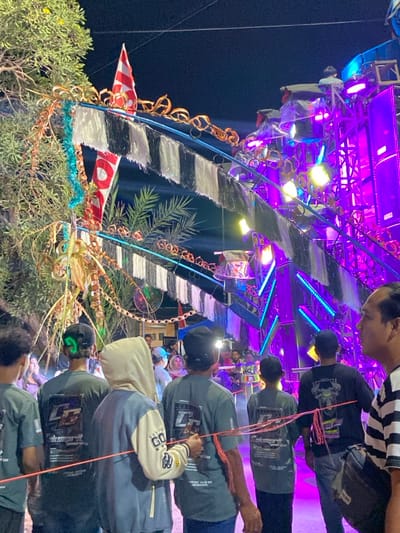 The image depicts a nighttime outdoor scene in Kedungbanteng, Indonesia, characterized by an elaborate, brightly lit setup and a gathering of individuals. In the background, a large, multi-tiered structure dominates the right side of the frame, heavily adorned with a sound system and numerous lights. These lights emit strong, vibrant purple, blue, and yellow illumination, creating a dynamic visual effect. The structure incorporates metal trussing, speakers, and various light fixtures.

Decorations are prominent, featuring an archway-like element decorated with gold-colored ornate scrolls at the top, below which hang horizontal black and white striped fabric streamers or fringe. Red and white triangular flags, consistent with the colors of the Indonesian flag, are positioned above and among these decorations. Green foliage, including palm fronds and tree branches, is integrated into the decorative scheme, particularly on the left side, alongside strands of turquoise tinsel and yellowish-brown fibrous material. A large tree with green leaves is visible on the far left.

In the mid-ground and foreground, a group of approximately seven individuals is observed mostly from behind, facing away from the camera and towards the illuminated structure. Most are wearing dark grey or greenish short-sleeved t-shirts featuring white graphic designs, such as "Gakuprit," on their backs. One individual in the center-left wears a light blue and cream-colored hooded jacket. Several wear dark baseball caps; one cap displays a bright green glowing element on its rear, and another has a headlamp attached to its front. A thin, reddish-orange string or cord extends across the backs of several individuals in the mid-foreground, appearing to connect them. On the far right, a person wearing a black and white striped shirt and a black fanny pack is partially visible, facing slightly right. The overall environment suggests an event or celebration under a dark night sky.