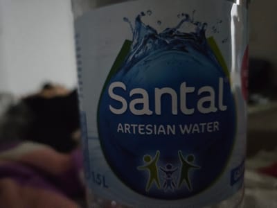 The image shows a close-up of a bottle of "Santal Artesian Water". The label is predominantly blue and features a stylized depiction of splashing water at the top. Below this, the brand name "Santal" is prominently displayed in white, followed by "ARTESIAN WATER" in smaller white text. At the bottom of the blue area, there are three silhouette figures of people, a larger one on the left and right with a smaller one in the middle, appearing to be a family unit. To the left of these figures, the text "1.5L" is visible. The background is out of focus and appears to be a domestic setting with muted colors and soft lighting. There is no information in the image that relates to the city of Yangikishak, Uzbekistan.