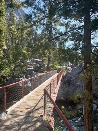 An eye-level, medium shot captures a wooden pedestrian bridge with red metal railings and chain-link fencing. The bridge spans a rocky ravine, with a body of water visible below and through the fencing. Lush green trees and large gray boulders populate the surrounding landscape. The sky above is clear and blue, with sunlight casting shadows across the bridge and the ground. The scene is set in a natural, outdoor environment, likely a park or hiking trail, with no people or discernible structures other than the bridge itself. The location is identified as Kanawyers, United States.