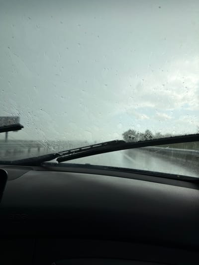 The image shows a view from inside a vehicle, likely a car, through the windshield. The windshield is covered in raindrops, indicating it is raining. A windshield wiper is partially visible, positioned vertically in the center of the frame. Outside, through the blurred and rain-streaked glass, the scene appears overcast and gray. The presence of a tree can be discerned in the distance.

The right side of the image is dominated by the interior of the vehicle, specifically the dashboard, which is dark gray or black and textured. An "SRS AIRBAG" label can be seen on the dashboard. The scene does not provide enough information to establish a location in Malko Tranovo, Bulgaria. No people, specific objects, actions, or interactions are visible.
