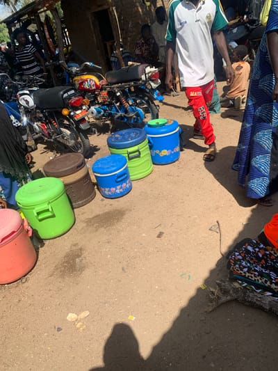 This image depicts a bustling scene outdoors, likely in Boi A, Nigeria, under the bright sun of a clear day. Several individuals are present, some of whom are engaged in activities related to a collection of objects. Prominently featured are several large plastic containers, in various colors including blue, green, and brown, some decorated with floral patterns. These containers appear to be either for sale or transport, as they are lined up on the ground.

Interspersed with the containers are motorcycles, suggesting a mode of transportation or perhaps a market setting where these items are sold. One person is seated on a motorcycle, dressed in a white shirt with green trim and red pants adorned with the number "57." Another individual, in an orange shirt, is leaning over, possibly interacting with the items or another person. Further back, a person in a black and white striped shirt is also near a motorcycle. The ground is a sandy, earthy surface, typical of an outdoor, possibly rural or semi-urban, environment. In the background, there are hints of structures and possibly more people and items, indicating a lively gathering.

The overall impression is one of commerce or daily life activity in a warm, sunny climate. The presence of people, motorcycles, and a variety of goods suggests a marketplace or a distribution point. The sunlight casts distinct shadows, confirming the time of day is likely midday. No overt emotions are discernible, but the arrangement of goods and people implies activity and interaction.