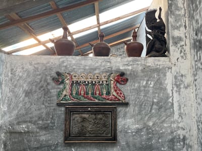 This image depicts a rustic interior scene in Karangkebagusan, Indonesia. The viewpoint is looking up at a shelf mounted on a textured concrete wall. On the shelf, from left to right, are three terracotta jugs with spouts, a dark wooden sculpture of a bird of prey perched on what appears to be another animal, and a partial view of another terracotta jug. Below the shelf, mounted on the wall, is a colorful wooden carving featuring multiple figures with heads resembling suns and bodies in red and green robes, flanked by dragon-like heads in red and green. Beneath this carving is a framed stone relief depicting a scene with figures. The ceiling above is constructed of corrugated metal with wooden beams, allowing sunlight to filter through gaps. The overall impression is one of traditional art and artifacts displayed in a simple, weathered setting.
