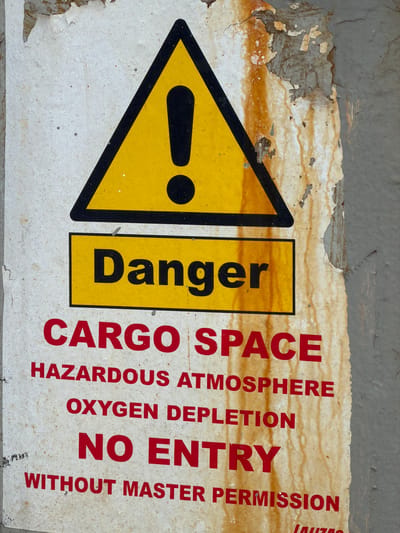 The image shows a weathered danger sign in Al Durra, Jordan. The sign is yellow with a black border and an exclamation mark inside a triangle, indicating a warning. Below the triangle, the word "Danger" is written in black on a yellow rectangle. The rest of the sign is white with red lettering. The text reads: "CARGO SPACE", "HAZARDOUS ATMOSPHERE", "OXYGEN DEPLETION", "NO ENTRY", and "WITHOUT MASTER PERMISSION". The sign is attached to a rough, textured wall that appears to be concrete or stone, with signs of rust and peeling paint. The overall condition of the sign suggests it has been exposed to the elements for a considerable time. There are no people or other objects visible in the immediate vicinity of the sign.