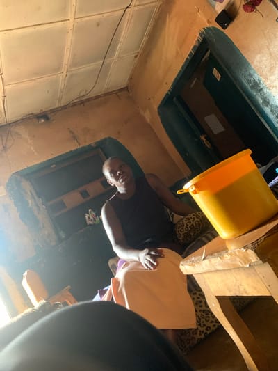 The image depicts an indoor scene, possibly a living room or common area, in Tabu, Nigeria. A woman is seated, wearing a dark sleeveless top and a light-colored skirt. She is looking towards the viewer. Beside her, on a wooden table, is a large, bright orange plastic bucket filled with a yellow substance. The room appears to be sparsely furnished with a sofa visible behind the woman, covered with a leopard-print fabric. The walls are textured and have a weathered appearance. The ceiling is composed of square panels. Natural light streams in from the left side of the frame, creating a bright highlight. The overall impression is one of a humble dwelling.