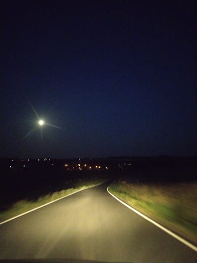 A nighttime view of a road in Veselá, Czechia, under a dark sky with a bright full moon. The road curves to the right, illuminated by headlights, and is bordered by grassy verges. In the distance, faint lights suggest a settlement or town. The overall scene depicts a tranquil, rural setting at night.