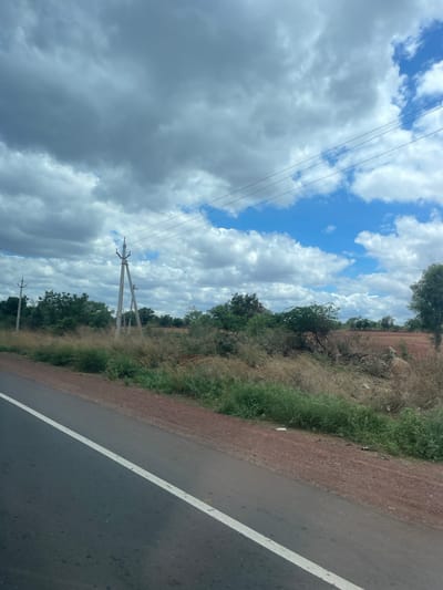 A roadside view captures a rural landscape under a cloudy sky. A grey asphalt road with a white line runs diagonally from the bottom left corner towards the right. The roadside verge is a mix of dry grass, brown soil, and sparse green vegetation. In the mid-ground, several tall, concrete utility poles with electrical wires strung between them are visible. Beyond the poles, there are scattered trees and shrubs, with some areas of red earth indicating cleared land or agricultural fields. The sky is partly cloudy, with large formations of grey and white clouds interspersed with patches of blue sky. The scene appears to be located in or around Sugali Tanda, India, given the presence of utility poles and vegetation typical of rural Indian landscapes. No people or vehicles are present in the image.